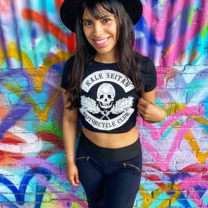 Woman with hat wearing kale seitan motorcycle shirt with skull and kale against graffiti wall