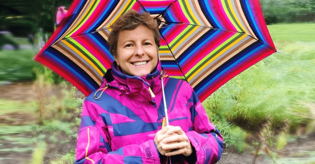 woman in pink rain jacket smiles with rainbow striped umbrella