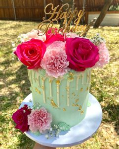 Green cake with assorted flowers, white drip frosting and a happy birthday topper